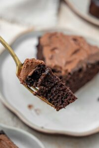 Close up of a fork holding a bite of chocolate cake with chocolate frosting. The rest of the slice is in the background.