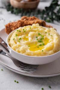 A white ceramic bowl filled with gold mashed potatoes with chives and melted butter on top. The bowl is on a ceramic plate with a piece of friend chicken next to the bowl. There are two forks tucked next to the bowl.