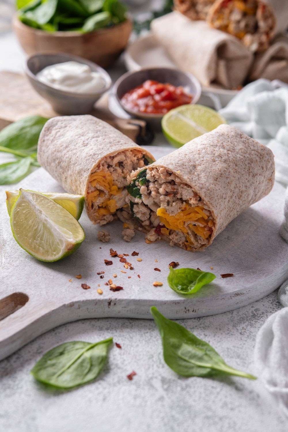 A high protein burrito sliced in half on a wooden server. The burrito has a whole wheat tortilla stuffed with ground turkey, cheese, egg whites, and spinach. There are spinach leaves and red pepper flakes sprinkled on the server.