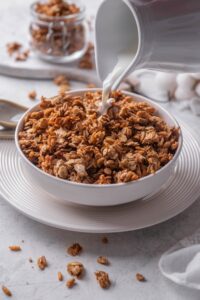 A white shallow bowl filled with granola with a pitcher of milk being poured on top of the granola.