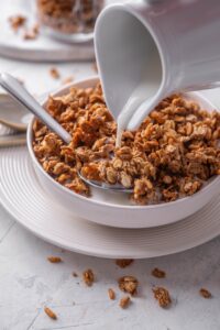 A white shallow bowl filled with granola with a pitcher of milk being poured on top of the granola. There is a spoon in the bowl and the bowl is on a white plate.
