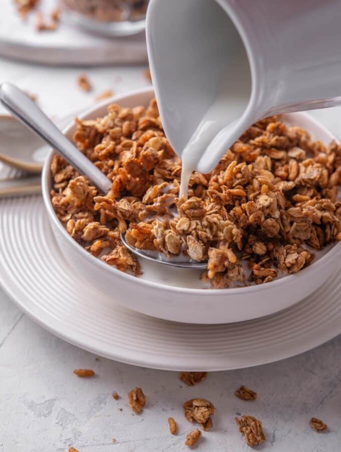 A white shallow bowl filled with granola with a pitcher of milk being poured on top of the granola. There is a spoon in the bowl and the bowl is on a white plate.