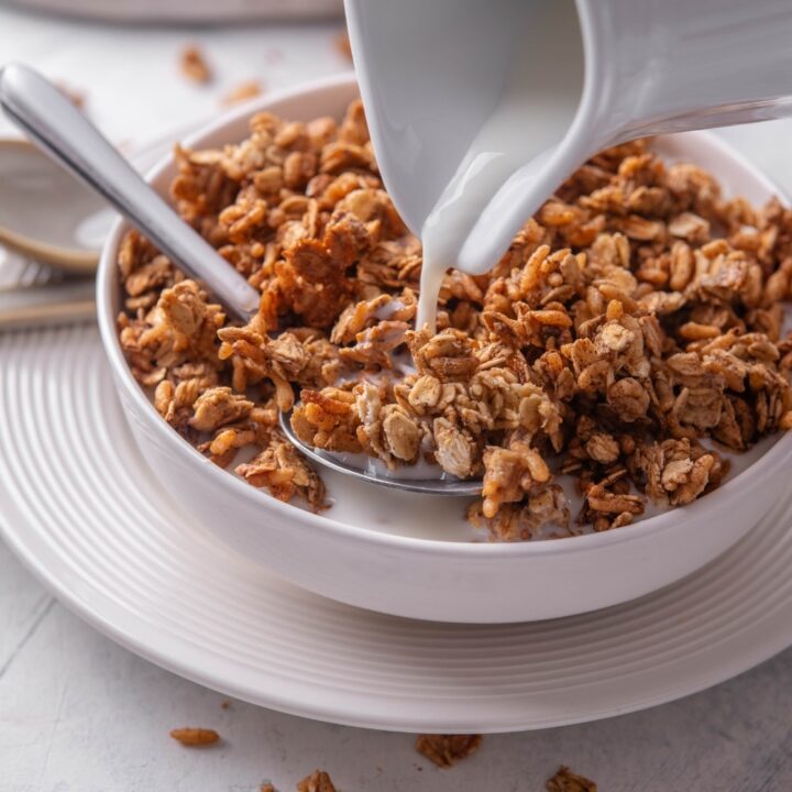 A white shallow bowl filled with granola with a pitcher of milk being poured on top of the granola. There is a spoon in the bowl and the bowl is on a white plate.