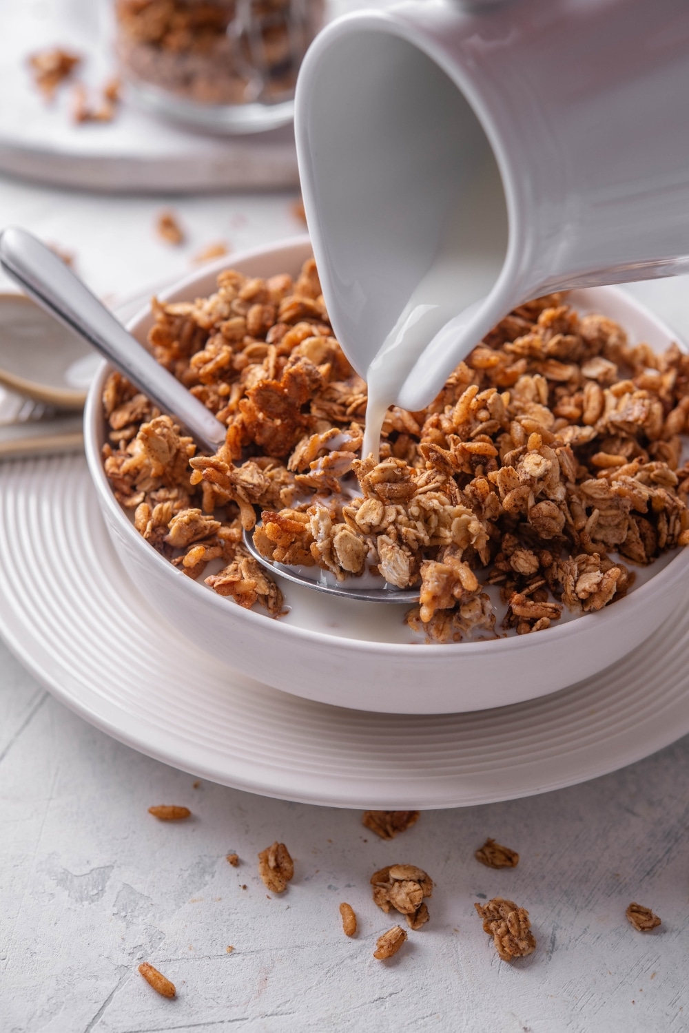 A white shallow bowl filled with granola with a pitcher of milk being poured on top of the granola. There is a spoon in the bowl and the bowl is on a white plate.