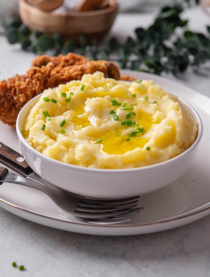 A white ceramic bowl filled with gold mashed potatoes with chives and melted butter on top. The bowl is on a ceramic plate with a piece of friend chicken next to the bowl. There are two forks tucked next to the bowl.