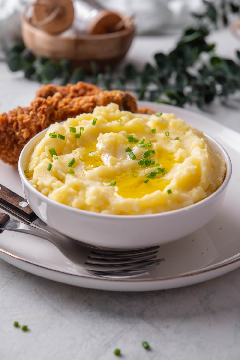 A white ceramic bowl filled with gold mashed potatoes with chives and melted butter on top. The bowl is on a ceramic plate with a piece of friend chicken next to the bowl. There are two forks tucked next to the bowl.