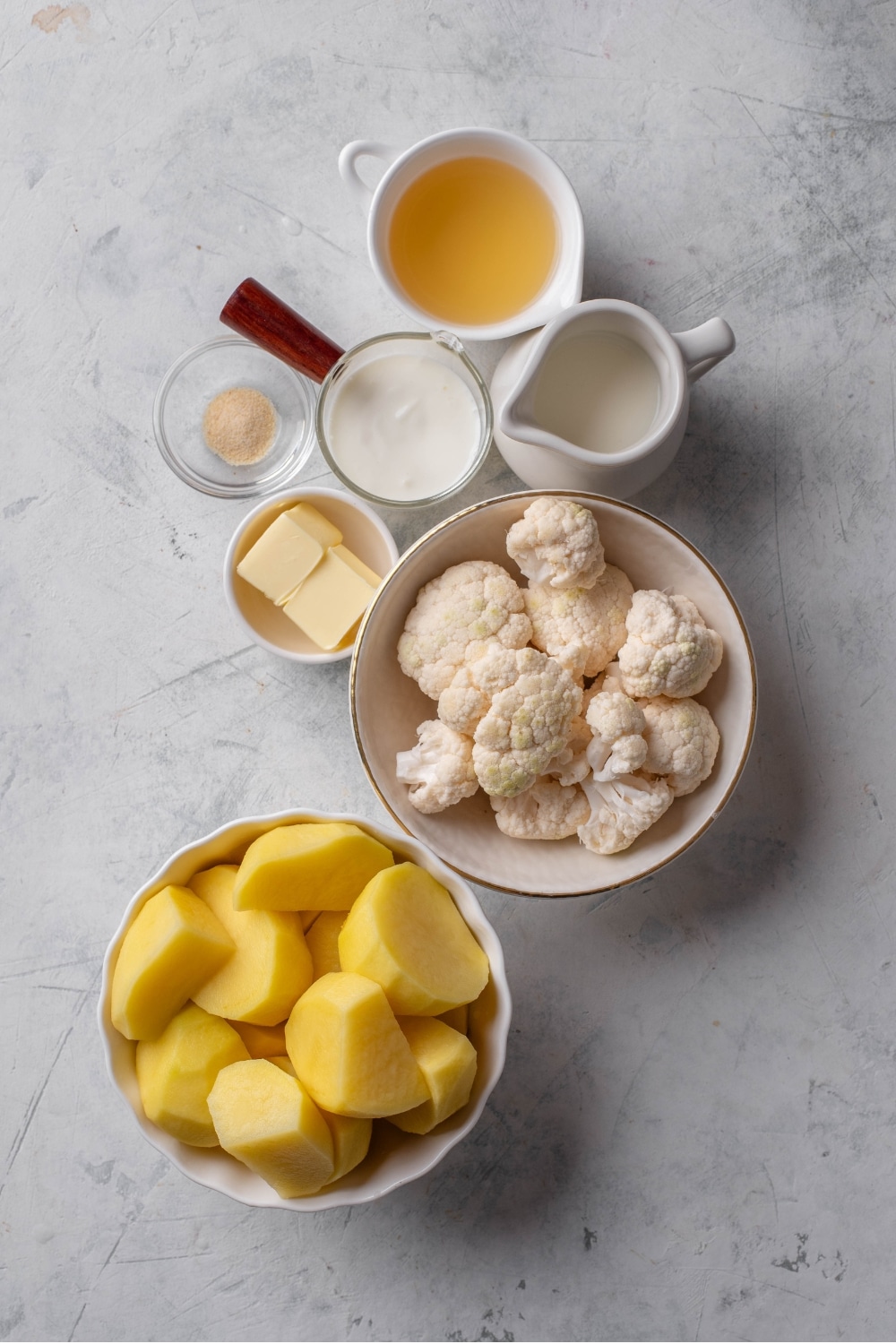 An overhead shot of several bowls of various shapes and sizes containing the ingredients to make healthy mashed potatoes including cubed gold potatoes, cauliflower, butter, greek yogurt, and chicken stock.