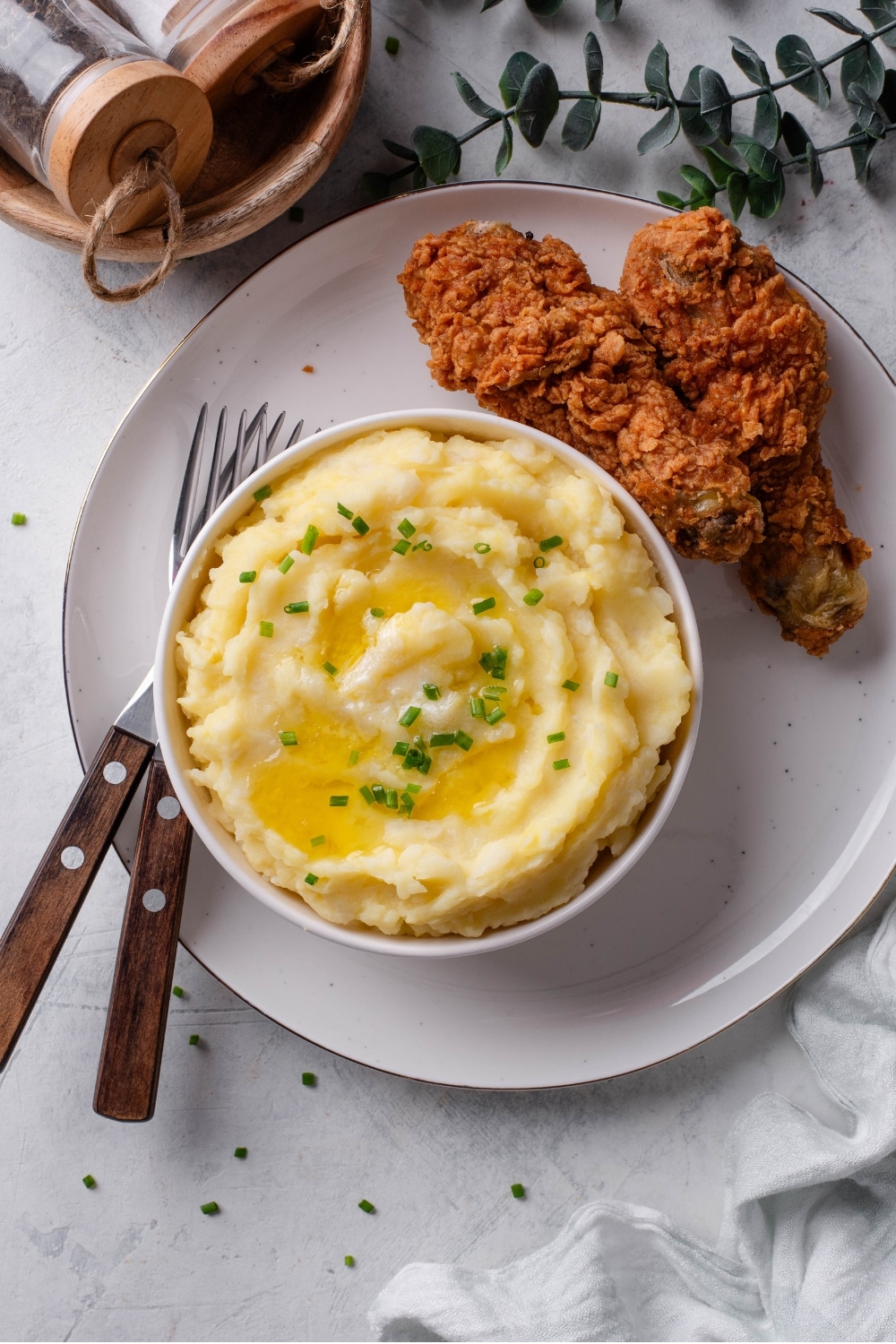 An overhead shot of a white ceramic bowl filled with gold mashed potatoes with chives and melted butter on top. The bowl is on a ceramic plate with two pieces of fried chicken next to it, and two forks on the other side of the bowl.