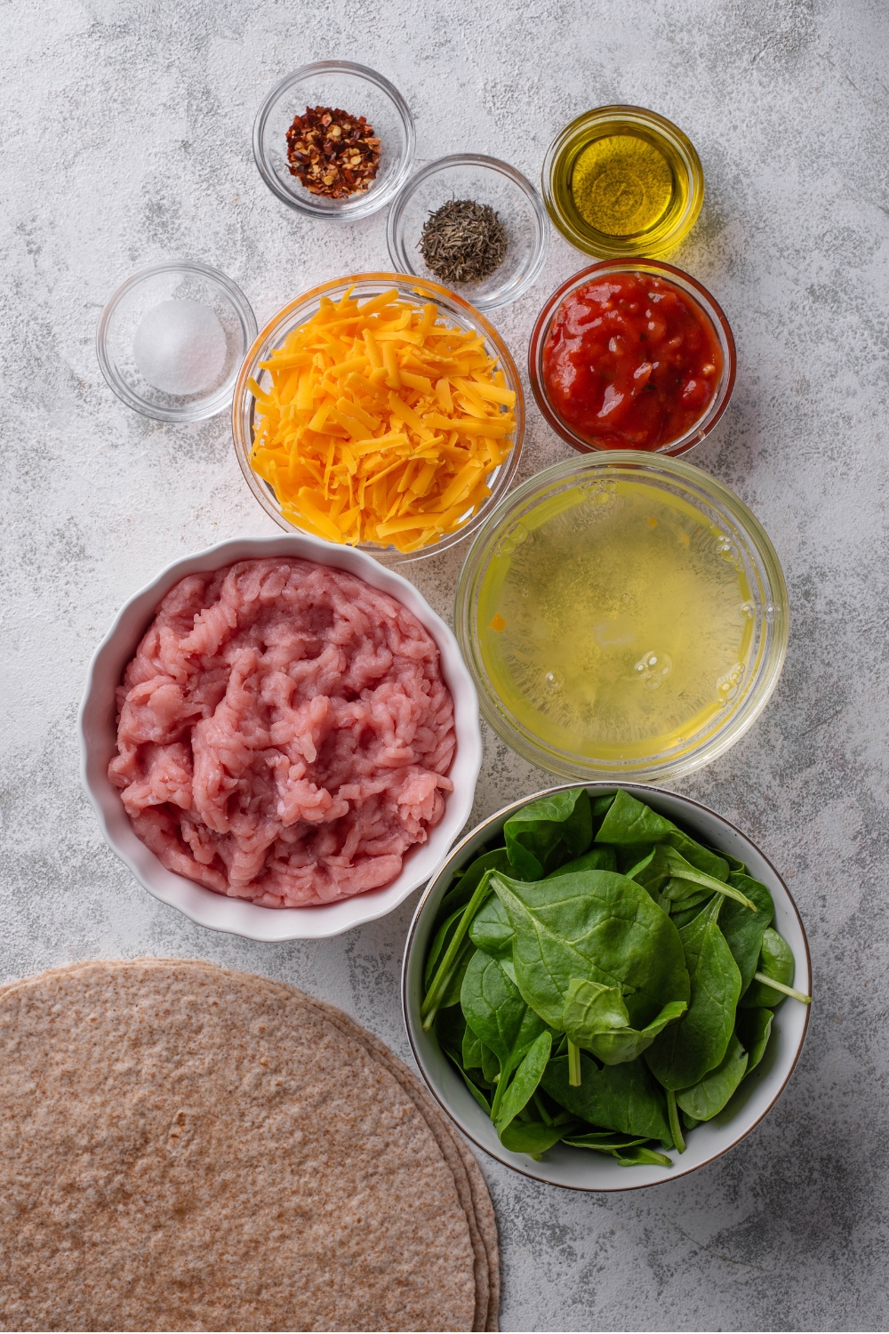 An overhead shot of several bowls of various sizes containing ingredients for high protein burritos including whole wheat tortillas, ground turkey, fresh spinach, egg whites, shredded cheese, red pepper flakes, and salsa.