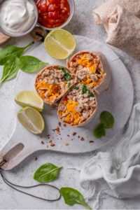 An overhead shot of three sliced high protein burritos stuffed with ground turkey, shredded cheese, spinach, and egg whites on a wooden server. There are lime wedges, spinach, and red pepper flakes sprinkled on the server with bowls of sour cream and salsa on the side.