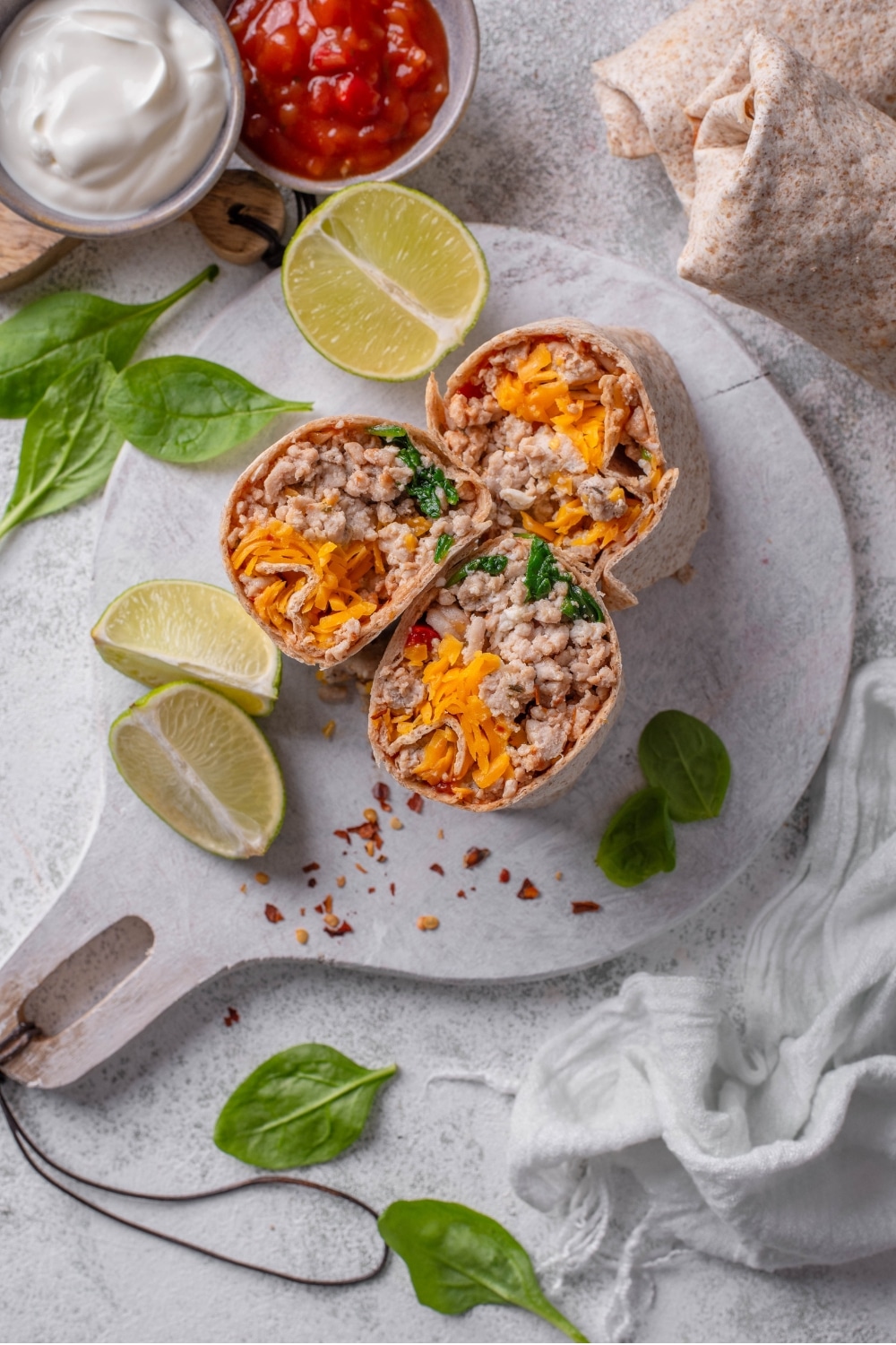An overhead shot of three sliced high protein burritos stuffed with ground turkey, shredded cheese, spinach, and egg whites on a wooden server. There are lime wedges, spinach, and red pepper flakes sprinkled on the server with bowls of sour cream and salsa on the side.