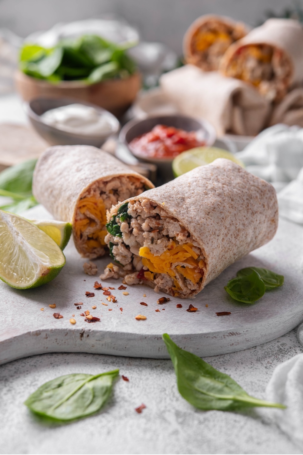 A high protein burrito sliced in half on a wooden server. The burrito has a whole wheat tortilla stuffed with ground turkey, cheese, egg whites, and spinach. There are spinach leaves and red pepper flakes sprinkled on the server.