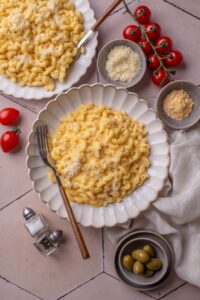 Overhead view of a plate of mac and cheese topped with cheese. There is a fork on the plate and a second plate of mac and cheese is next to it. The plates are surrounded with small bowls of other ingredients, olives, and a bunch of cherry tomatoes.