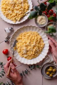 Overhead view of a plate of mac and cheese topped with cheese. The plate is being held with one hand and the other hand is holding a fork. A second plate of mac and cheese is next to it.