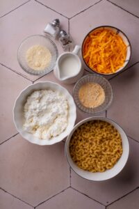 An assortment of ingredients including a pitcher of milk, mini salt and pepper shakers, and bowls of shredded cheese, dried pasta, cottage cheese, and seasonings.