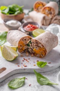 A high protein burrito sliced in half on a wooden server. The burrito has a whole wheat tortilla stuffed with ground turkey, cheese, egg whites, and spinach. There are spinach leaves and red pepper flakes sprinkled on the server.