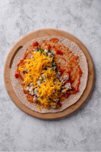An overhead shot of a whole wheat tortilla with salsa, ground turkey, spinach, and shredded cheese layered on top. The tortilla is on a wooden tray.