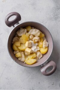 An overhead shot of a stock pot filled with cooked cubed potatoes, cauliflower, pats of butter, and greek yogurt on a marble countertop.