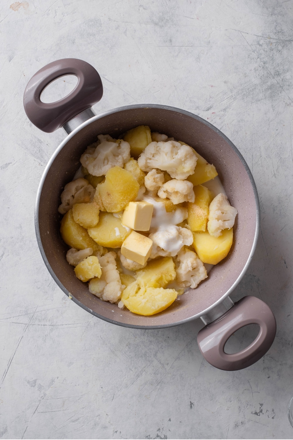 An overhead shot of a stock pot filled with cooked cubed potatoes, cauliflower, pats of butter, and greek yogurt on a marble countertop.