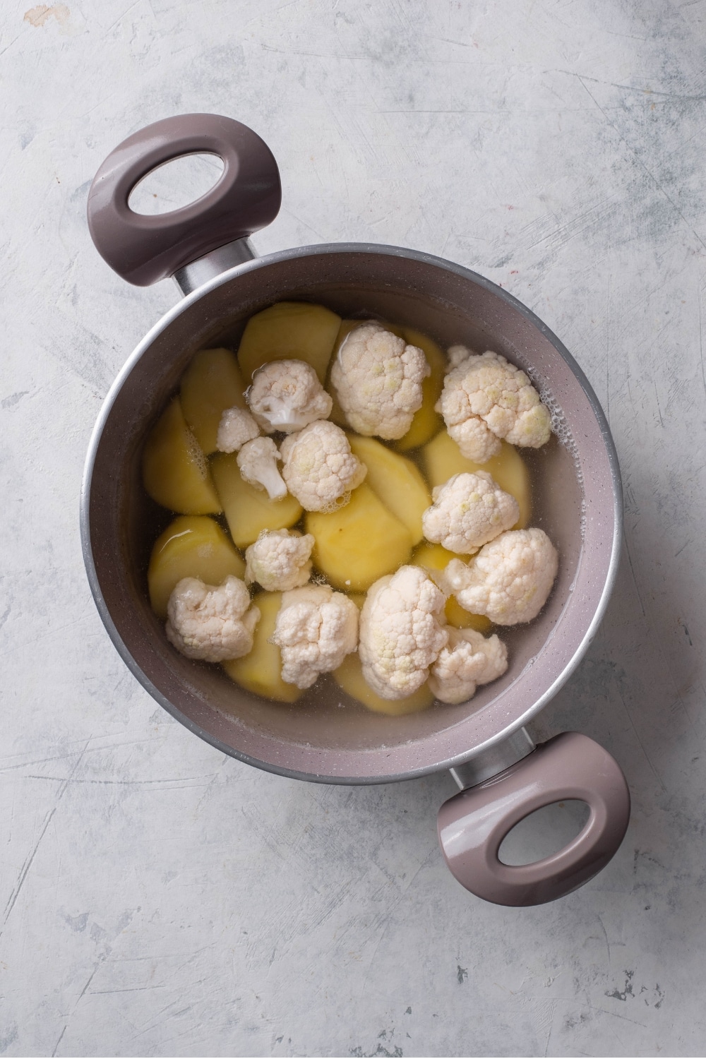 An overhead shot of a stock pot filled with water, cubed potatoes, and cauliflower on a marble counter.