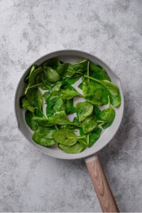 An overhead shot of a ceramic skillet with cooked spinach on a marble countertop.