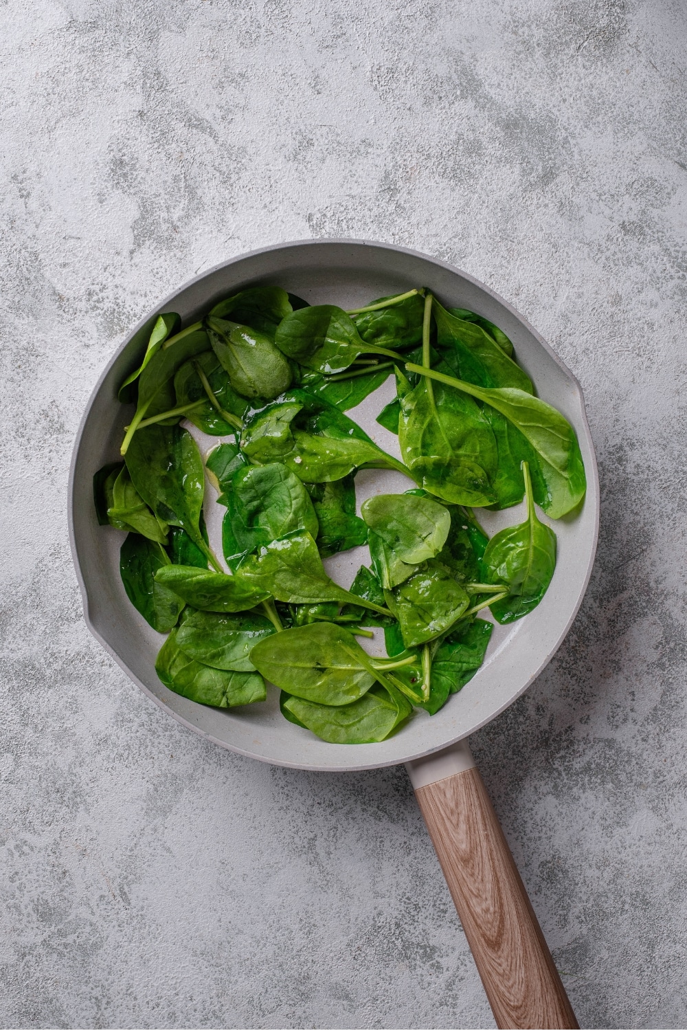 An overhead shot of a ceramic skillet with cooked spinach on a marble countertop.