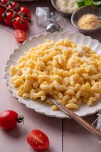 A plate of mac and cheese with a fork on the plate. The plate is surrounded by an assortment of ingredients and cherry tomatoes spilled onto the counter.