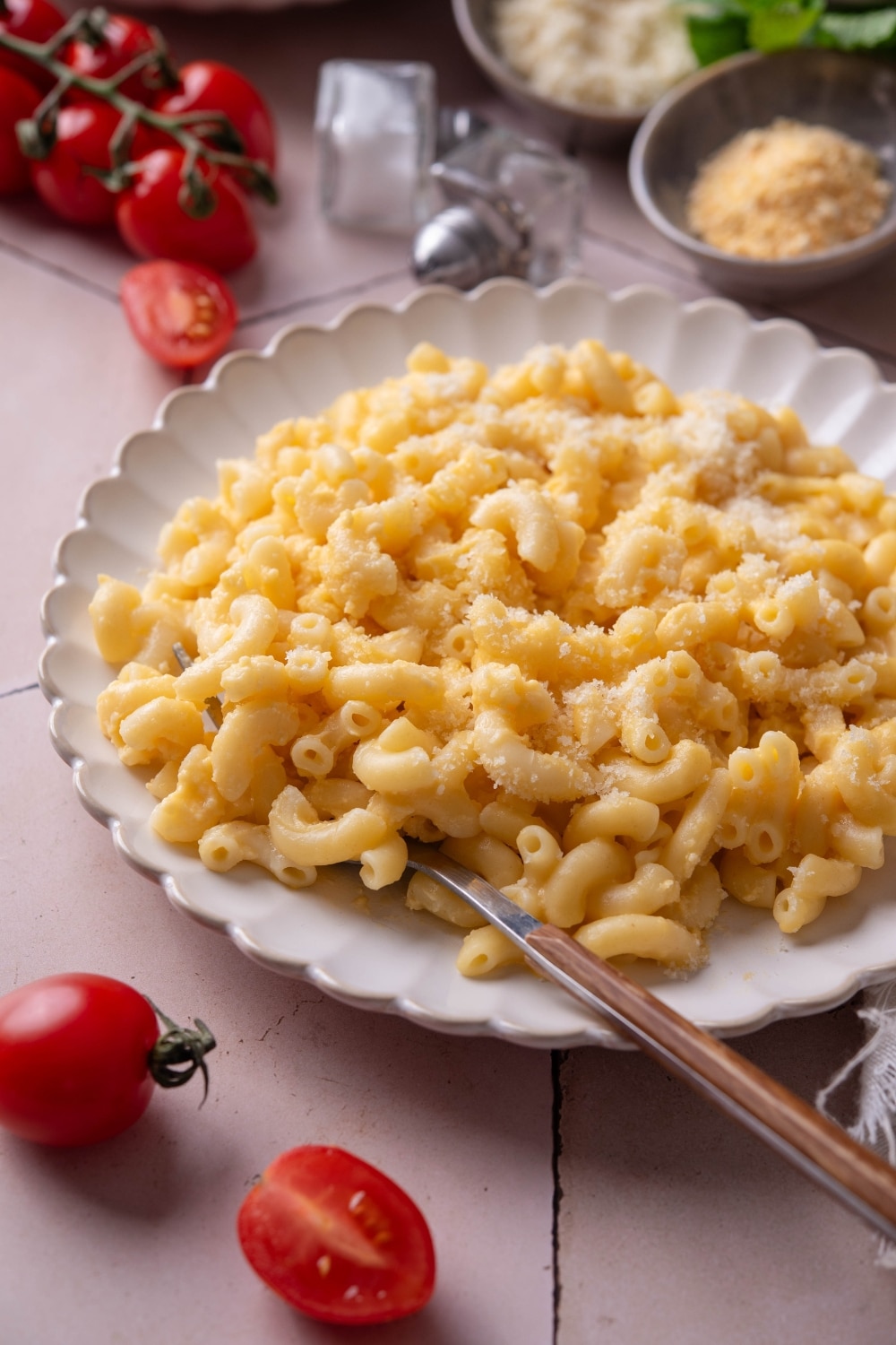 A plate of mac and cheese with a fork on the plate. The plate is surrounded by an assortment of ingredients and cherry tomatoes spilled onto the counter.