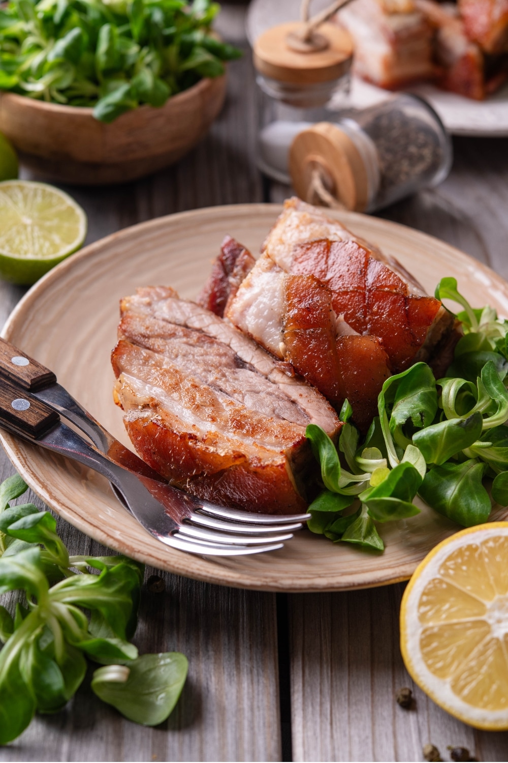 A tan ceramic plate with two pieces of sliced pork belly with a small side of greens. There are two forks on the plate and a halved lemon in front of the plate.