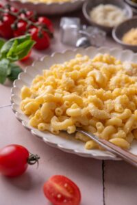 A plate of mac and cheese with a fork on the plate. The plate is surrounded by an assortment of ingredients and cherry tomatoes spilled onto the counter.