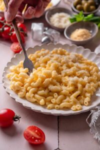 A plate of mac and cheese with a fork being stuck into the plate. The plate is surrounded by an assortment of ingredients and cherry tomatoes spilled onto the counter.