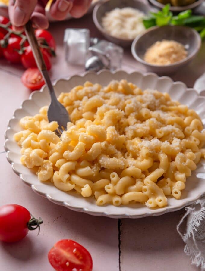 A plate of mac and cheese with a fork being stuck into the plate. The plate is surrounded by an assortment of ingredients and cherry tomatoes spilled onto the counter.