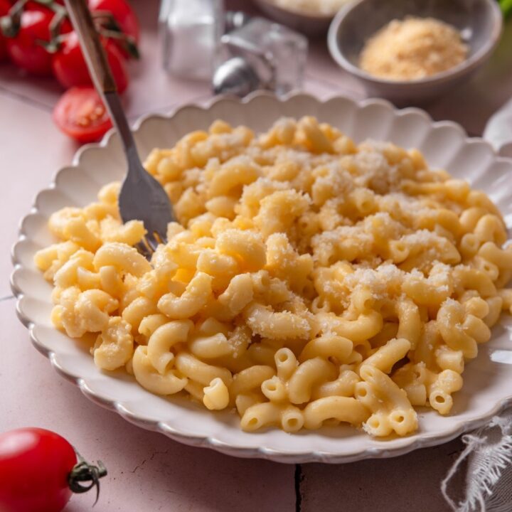 A plate of mac and cheese with a fork being stuck into the plate. The plate is surrounded by an assortment of ingredients and cherry tomatoes spilled onto the counter.