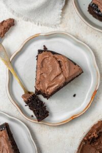 A slice of chocolate cake covered in chocolate frosting on a plate with a fork holding a bite of the cake.