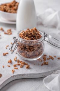 A clear jar filled with granola with pieces of granola spilling over the jar onto the counter.