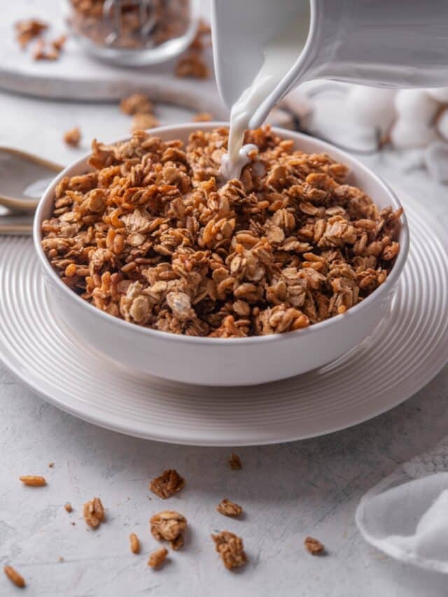 A white shallow bowl filled with granola with a pitcher of milk being poured on top of the granola.