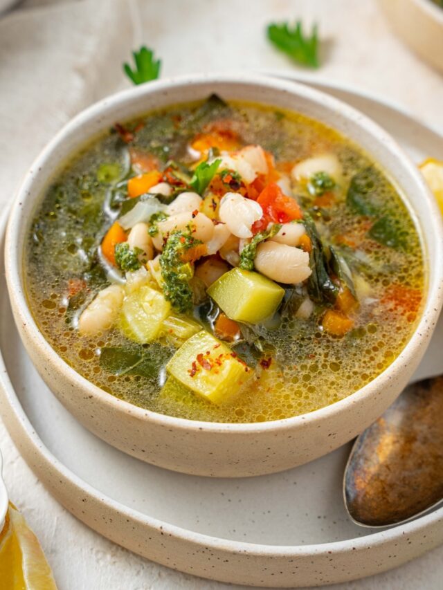 Overhead view of a white bowl filled with vegetable soup with pieces of carrot, tomato, kale, zucchini, and white beans. The bowl is on a plate with a spoon.