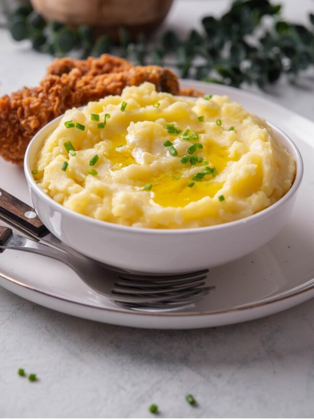 A white ceramic bowl filled with gold mashed potatoes with chives and melted butter on top. The bowl is on a ceramic plate with a piece of friend chicken next to the bowl. There are two forks tucked next to the bowl.