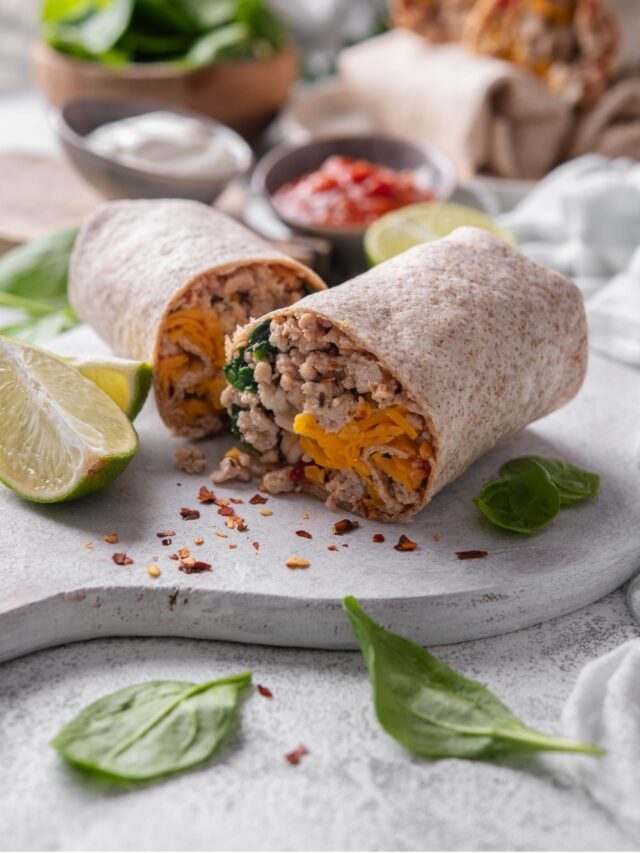 A high protein burrito sliced in half on a wooden server. The burrito has a whole wheat tortilla stuffed with ground turkey, cheese, egg whites, and spinach. There are spinach leaves and red pepper flakes sprinkled on the server.