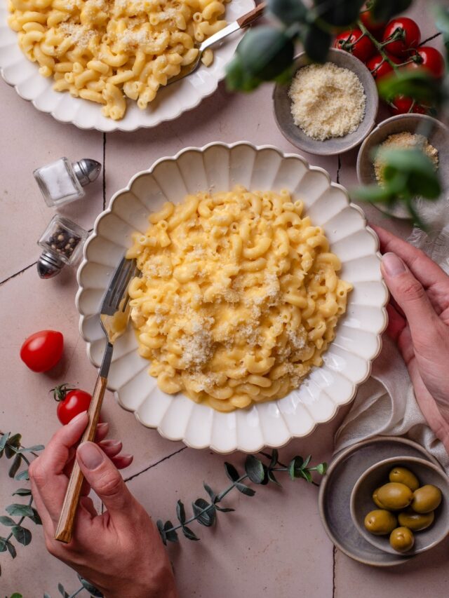 Overhead view of a plate of mac and cheese topped with cheese. The plate is being held with one hand and the other hand is holding a fork. A second plate of mac and cheese is next to it.