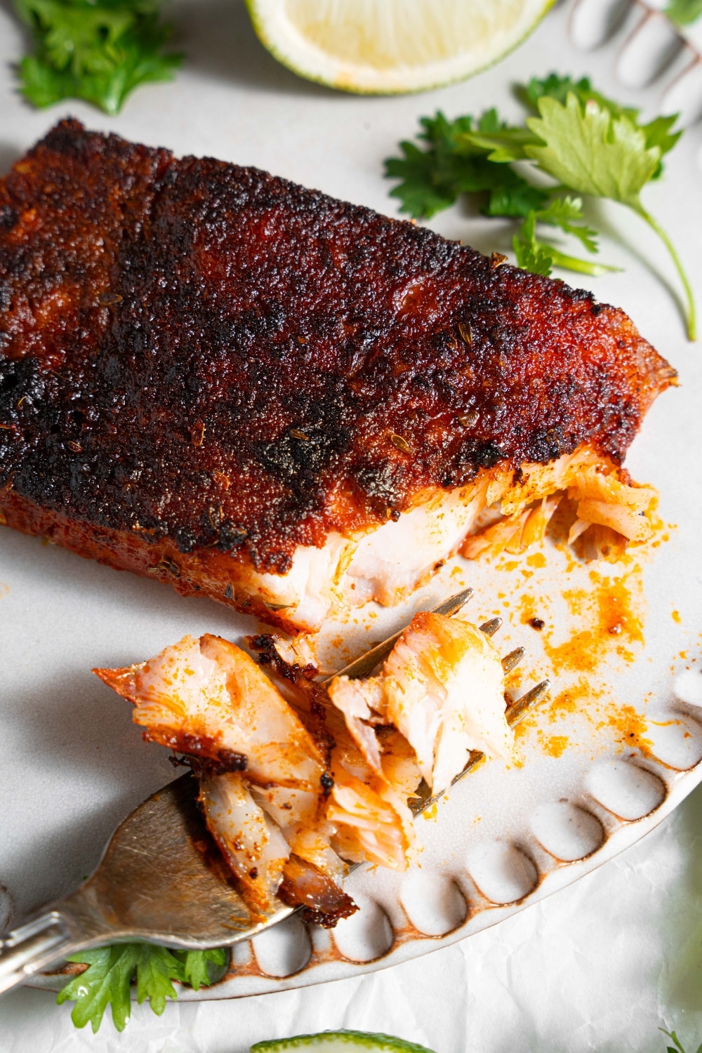 A close up of blackened mahi mahi on a ceramic plate with a fork taking a bite of fish. The plate is garnished with parsley and lime.