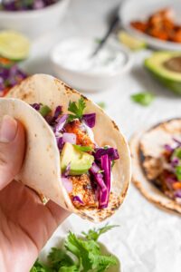 A close up of a hand holding a mahi mahi taco with an avocado slaw topping. There is an additional plate of tacos and bowls of ingredients in the background.