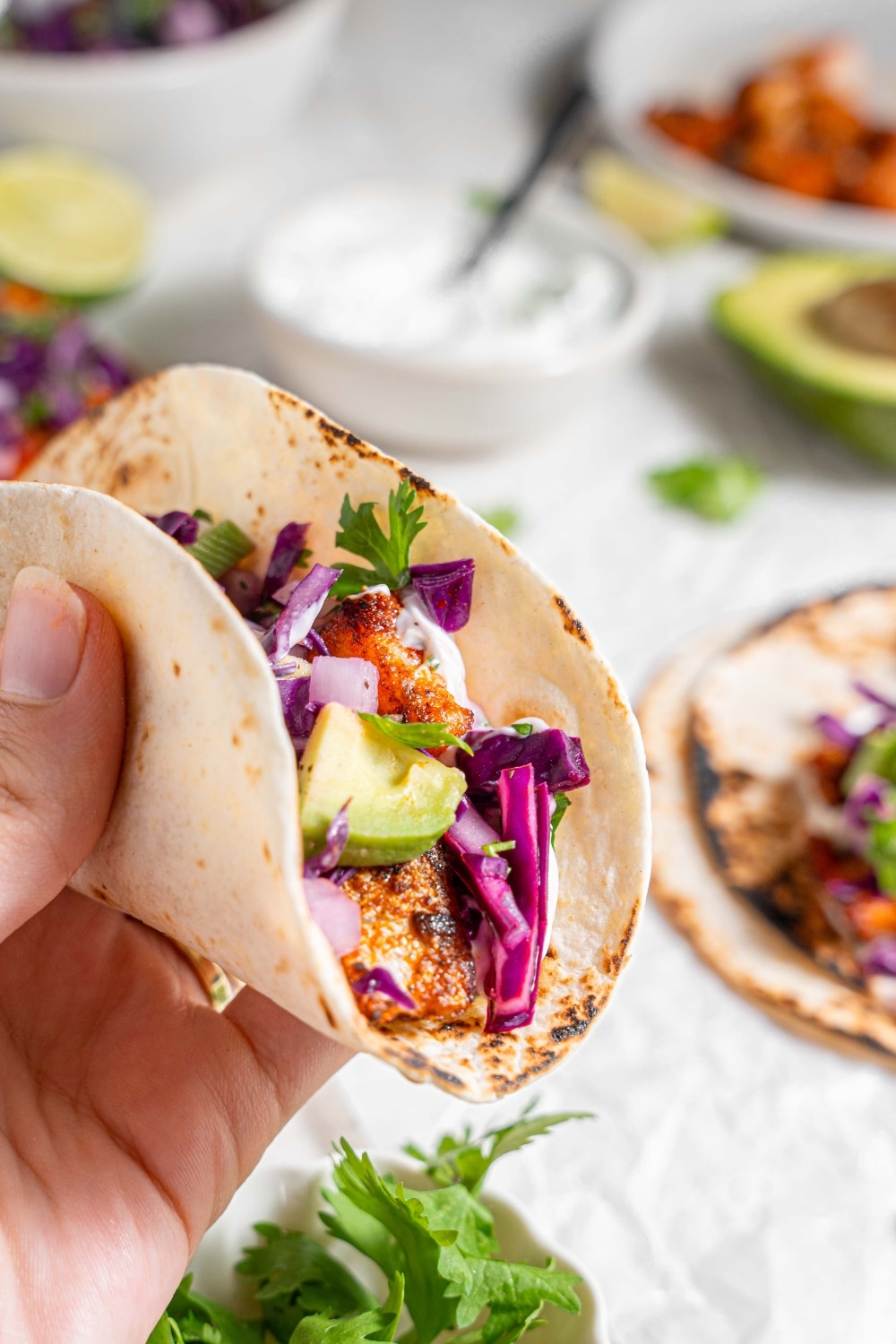 A close up of a hand holding a mahi mahi taco with an avocado slaw topping. There is an additional plate of tacos and bowls of ingredients in the background.