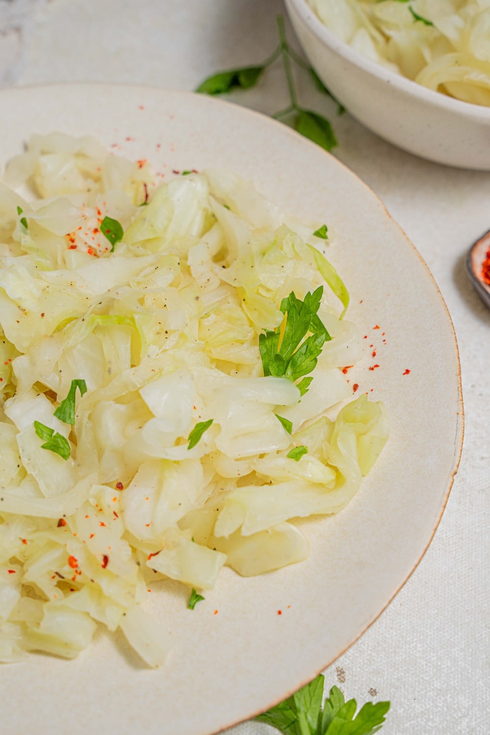 A plate of steamed cabbage on a marble counter with an additional bowl of steamed cabbage off to the side. The plate and counter are garnished with fresh parsley.