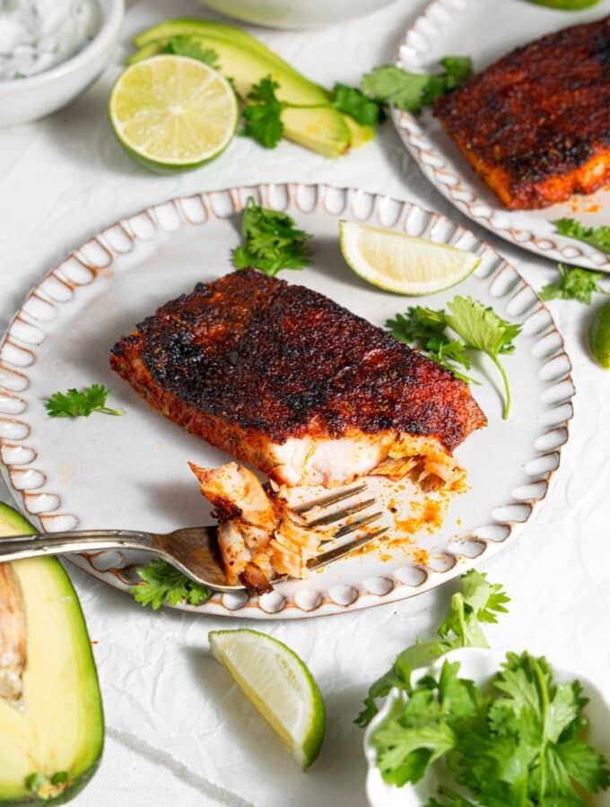 A ceramic plate with blackened mahi mahi with a fork taking a bite of fish. The plate is garnished with parsley and lime. There is a second plate of blackened mahi mahi and additional garnishes next to the plate.