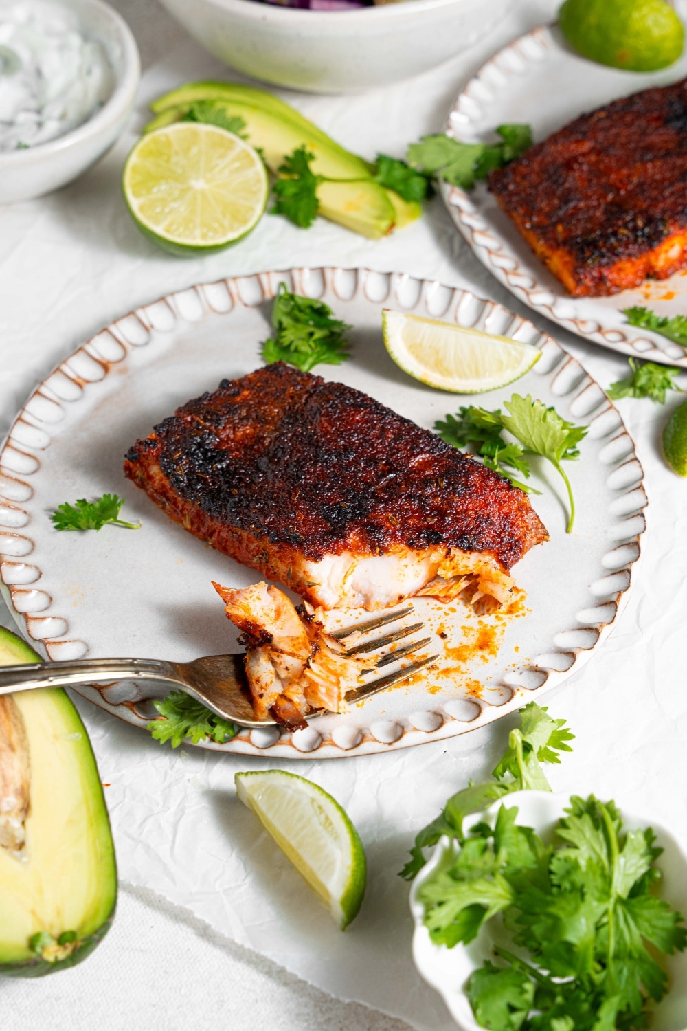 A ceramic plate with blackened mahi mahi with a fork taking a bite of fish. The plate is garnished with parsley and lime. There is a second plate of blackened mahi mahi and additional garnishes next to the plate.