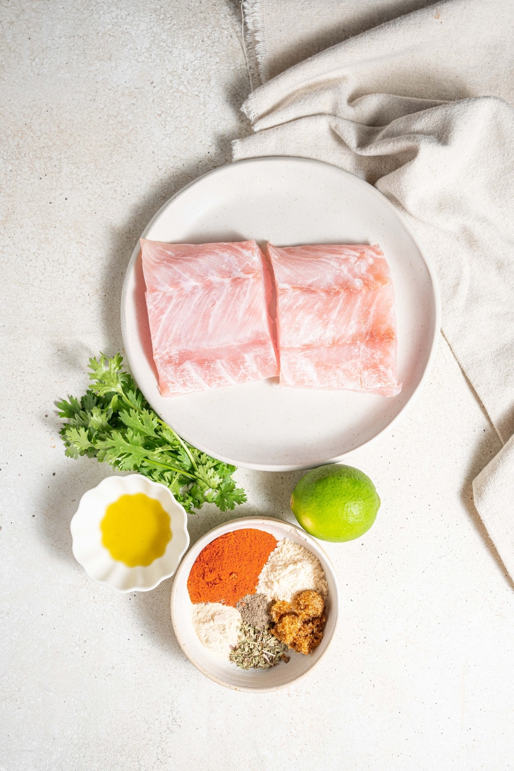 An overhead shot of several plates and bowls of various sizes containing ingredients to make blackened mahi mahi including a plate with two mahi mahi fillets, and bowls filled with olive oil, and cajun seasonings.