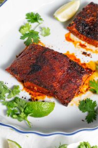A piece of cooked blackened mahi mahi on a baking dish garnished with sprigs of parsley and a lime wedge. There is a second piece of cooked blackened mahi mahi in the baking dish.