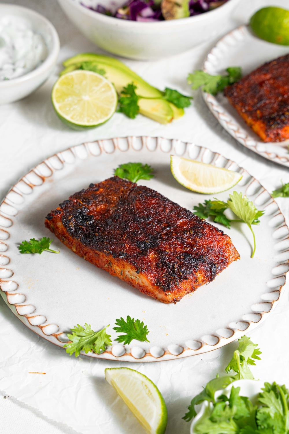 A ceramic plate with blackened mahi mahi on a marble counter. The plate is garnished with parsley and lime. There is a second plate of blackened mahi mahi and additional garnishes next to the plate.