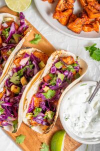 A wooden server with three mahi mahi tacos next to a bowl of sour cream sauce. Everything is garnished with cilantro and there is an extra plate of fried mahi mahi on the side.